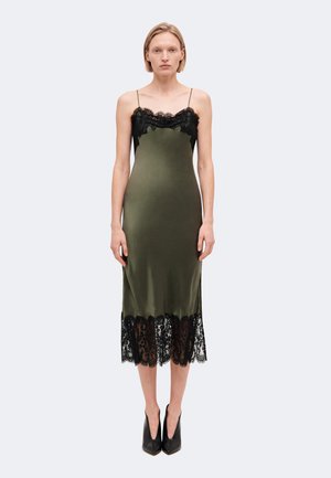 Femme portant une robe nuisette en satin vert olive avec une bordure en dentelle noire au niveau du col et de l'ourlet, associée à des escarpins noirs pointus, debout.