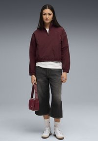 Kvinde står iført en maroon zip-up jakke, hvid skjorte, sorte cropped jeans, hvide sokker, hvide sneakers og holder en maroon håndtaske.