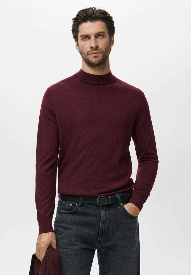 Hombre vestido con un suéter de cuello alto burdeos de mangas largas y jeans negros, sosteniendo una prenda burdeos a juego en su mano izquierda.