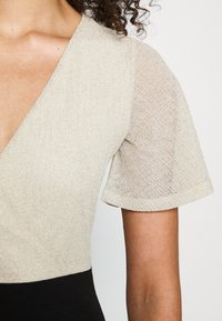 Haut en tricot beige avec un décolleté en V profond et des manches courtes en transparence ; tissu texturé avec des fils métalliques ; bas noir ajusté.