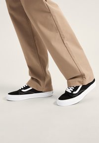 Sneakers in suede neri con dettagli e lacci bianchi, caratterizzati da un design low-top e una spessa suola in gomma bianca, abbinati a pantaloni khaki.
