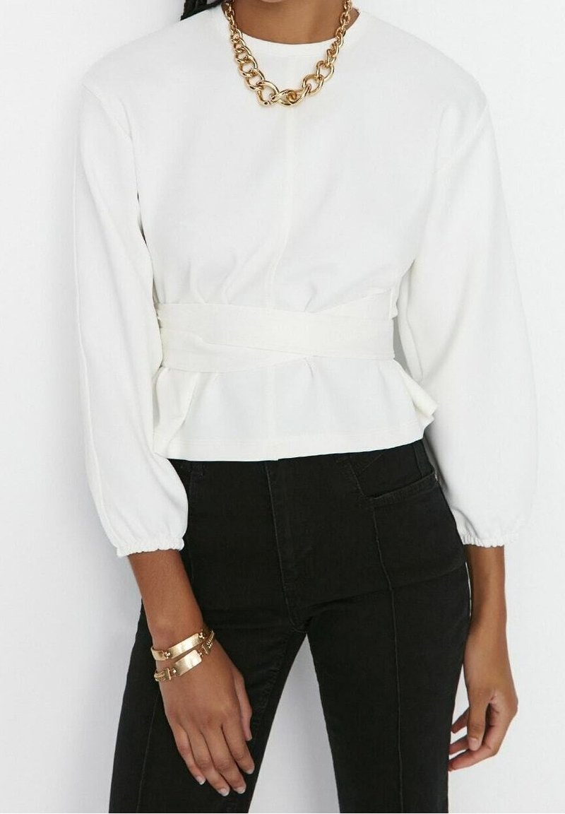 Femme portant un haut blanc à manches longues avec une large ceinture à la taille, un pantalon noir, un collier chaîne épaisse dorée et des bracelets en or au poignet gauche.