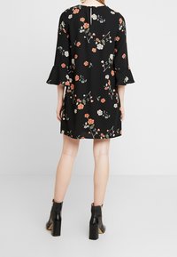 Žena stojící, oblečená v černých květinových šatech s 3/4 zvonovými rukávy a černými kotníkovými botami, zobrazená zezadu na jednobarevném šedém pozadí.