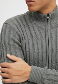 Grauer gestrickter Pullover mit hohem Kragen, der ein strukturiertes Kabelmuster aufweist. Reißverschlussdetail am Hals und gerippte Bündchen.
