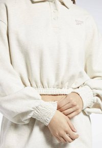 Personne portant un sweat-shirt court de couleur crème avec une taille et des poignets élastiques, attachant les manches devant avec les mains.