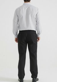 Homme portant une chemise à manches longues blanche à motifs, un pantalon de costume noir et des chaussures de costume noires, se tenant de dos sur fond uni.