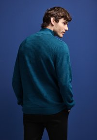 Pull en rib à col montant de couleur teal avec un design texturé, associé à un pantalon foncé sur fond bleu.