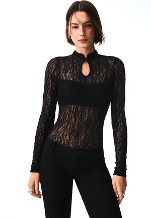 Donna che indossa un top nero in pizzo a manica lunga con scollo a goccia, abbinato a pantaloni neri aderenti su sfondo bianco.