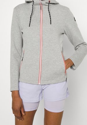 Person wearing light gray zip-up hoodie with pink zipper and black drawstrings, paired with light purple layered shorts, one hand in pocket.