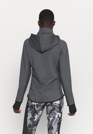 Person iført en mørkegrå hættetrøje og sorte, hvide og grå mønstrede leggings, vist bagfra mod en ensfarvet baggrund.