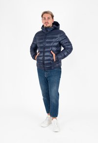 Marineblauwe pufferjack met capuchon, voorzien van een gestikte ontwerp en ritssluiting, gecombineerd met blauwe jeans en witte sneakers.