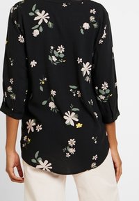 Blouse noire à manches ¾, avec un motif floral rose et jaune, ourlet arrondi et coupe décontractée. Tissu doux et léger.