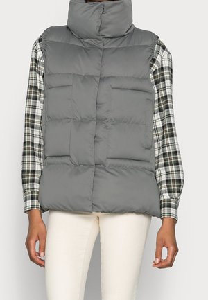 Veste matelassée grise portée sur une chemise à carreaux noir et blanc à manches longues, associée à un pantalon écru, montrée sur une personne debout.