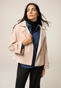 Lichtroze jack met gestructureerde revers, zijrits en manchettdetails, gelaagd over een blauwe spijkerbloes en een zwarte coltrui.