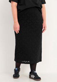 Schwarzer Midirock aus Spitzenstoff mit einem texturierten Blumenmuster. Kombiniert mit schwarzen Sneakers und blickdichten schwarzen Strumpfhosen.