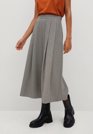 Mujer con una falda midi gris a cuadros de talle alto con pliegues, botas negras gruesas y una camiseta sin mangas naranja.