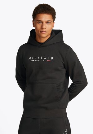 Jeune homme portant un sweat à capuche noir avec le texte "HILFIGER NEW YORK" et un pantalon noir assorti, debout avec les mains dans le dos.