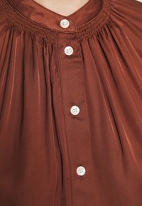 Bruine, knoopsluiting blouse met een geplooid ontwerp en gesmokte halslijn. Voorzien van witte knopen en een gladde, geweven stof.