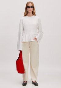 Witte lange blouse met ruches aan de onderkant, crèmekleurige wijde broek, en een rode schoudertas. Zwarte instappers maken de outfit compleet.
