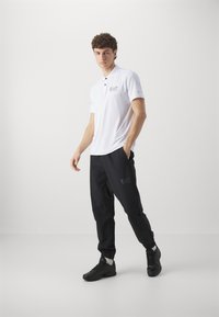 White polo shirt with short sleeves, featuring logo, paired with tapered black pants with green logo, and black athletic shoes. Simple design.