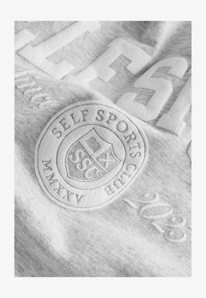 Siva tkanina dukserice s teksturiranim natpisima, koja sadrži logo "SELF SPORTS CLUB", godinu "2023" i utisnut okrugli grb.