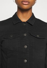 Giacca di jeans nera con colletto, bottoni a pressione argentati e due tasche a pattina sul petto. Il tessuto ha una consistenza liscia.