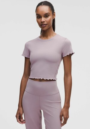 Kvinde stående iført en matchende lys lilla ribbet cropped top og højtaljede bukser med bølgede kanter, mod en ensfarvet lys baggrund.