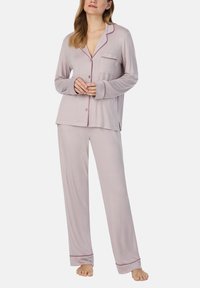 Vrouw in een lichtroze, lang mouw, knoopdetails slaapshirt en bijpassende losse broek, staand op blote voeten tegen een effen achtergrond.