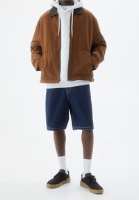Brązowa kanwasowa kurtka z szarym kapturem pod spodem, zestawiona z niebieskimi dżinsowymi szortami i czarnymi trampkami z gumową podeszwą.