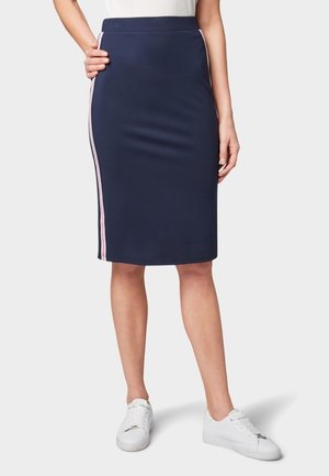 Femme portant une jupe bleu marine longueur genou avec des bandes latérales roses, un haut blanc et des baskets blanches, debout devant un fond uni.