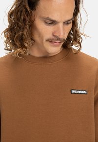 Sudadera marrón con cuello de canalé, que presenta un parche de logo bordado en negro en el lado izquierdo. Hecha de una suave tela texturizada.
