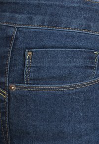 Dark blue denim jeans featuring a small front pocket, visible stitching in a lighter shade, and metal rivets near the pocket edge.