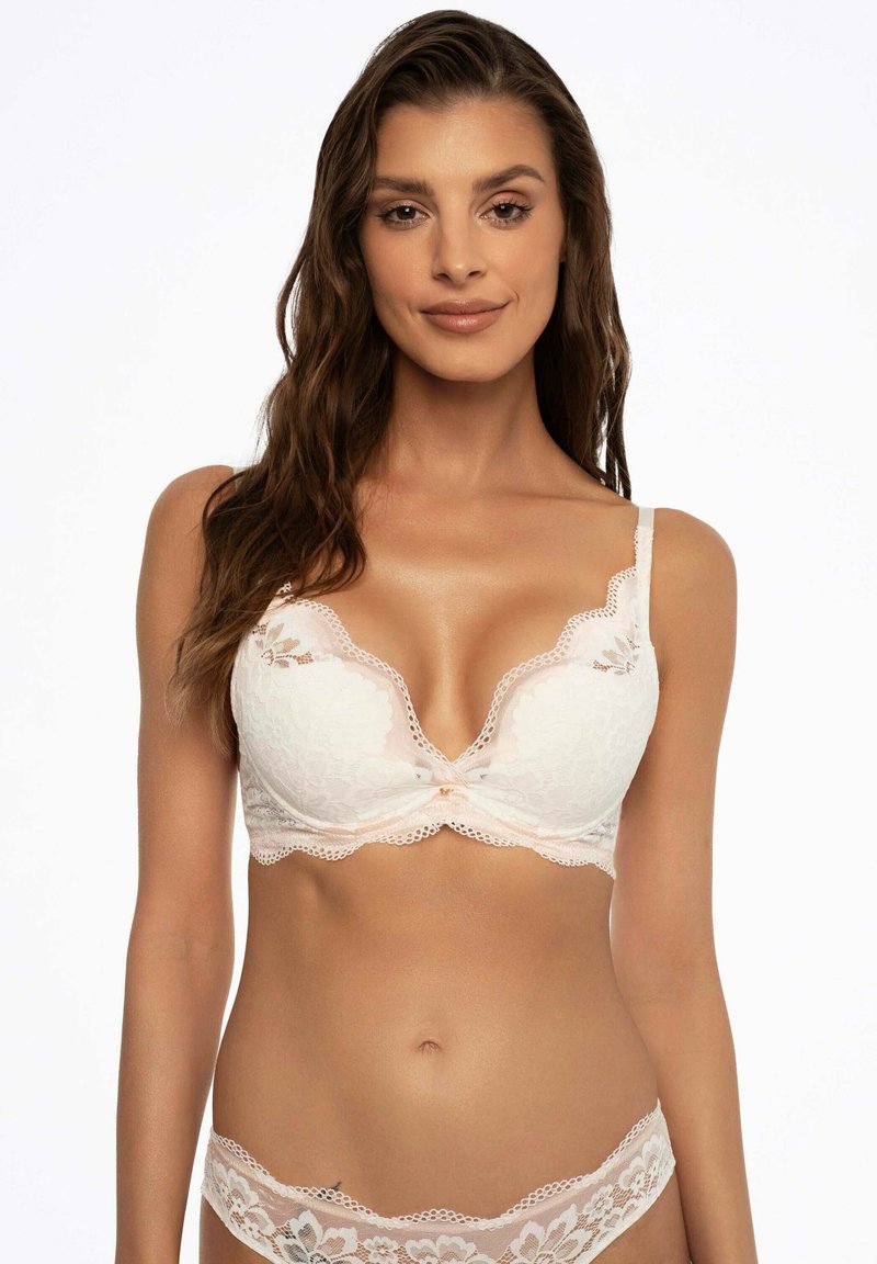 Kobieta z długimi brązowymi włosami, ubrana w biały koronkowy biustonosz i pasujące koronkowe majtki na jednolitym białym tle.