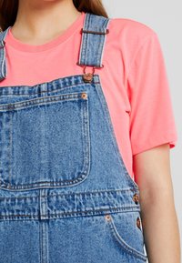 Denim haalarit keskisinisessä värissä, säädettävillä olkaimilla, kuparinvärisillä metalliosilla ja etutaskulla, yhdistettynä kirkkaan vaaleanpunaiseen lyhythihaiseen t-paitaan.