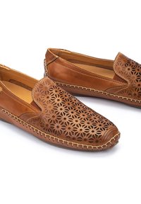 Chaussures à enfiler en cuir marron avec un motif floral découpé sur la partie avant, accentuées de coutures beiges le long de la semelle. Texture lisse.