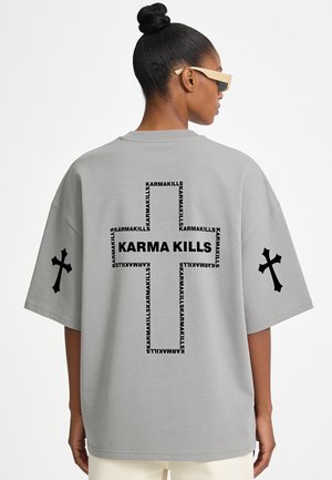 Frau mit Hochsteckfrisur, die beige Sonnenbrillen und ein graues übergroßes T-Shirt mit schwarzen Kreuzen und dem Text "KARMA KILLS" auf Rücken und Ärmeln trägt.