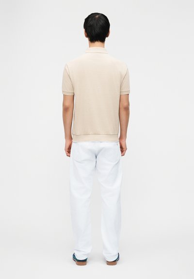 Άνδρας που στέκεται με την πλάτη στραμμένη, φορώντας μπεζ κοντομάνικη πόλο μπλούζα, λευκό παντελόνι και σκούρα παπούτσια, μπροστά σε απλό ανοιχτό φόντο.