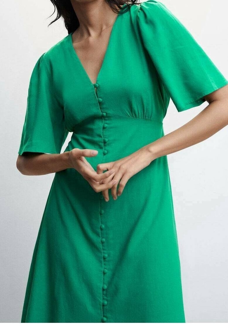 Femme portant une robe vert vif à col en V avec des manches courtes et des boutons recouverts de tissu à l'avant, mains légèrement jointes.
