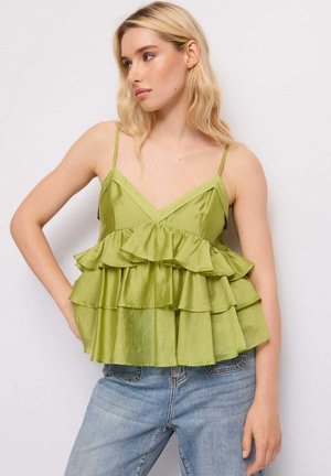 Donna con capelli biondi che indossa un top smanicato verde lime con ruches e jeans azzurri, in piedi davanti a uno sfondo bianco neutro.