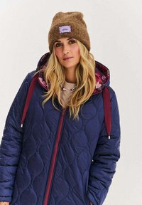 Granatowa pikowana kurtka z bordowymi akcentami i kwiatową podszewką, w połączeniu z brązową dzianą czapką z jasnofioletową łatą.