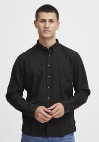 Camisa negra de manga larga, confeccionada en una tela texturada, con collar tipo spread. Presenta botones en tono dorado y un dobladillo recto.