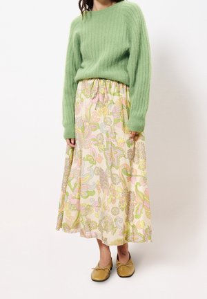 Vrouw met een lichtgroene, zachte trui, pastelkleurige midi-rok met bloemenprint en mosterdgele ballerina's, staand tegen een effen witte achtergrond.
