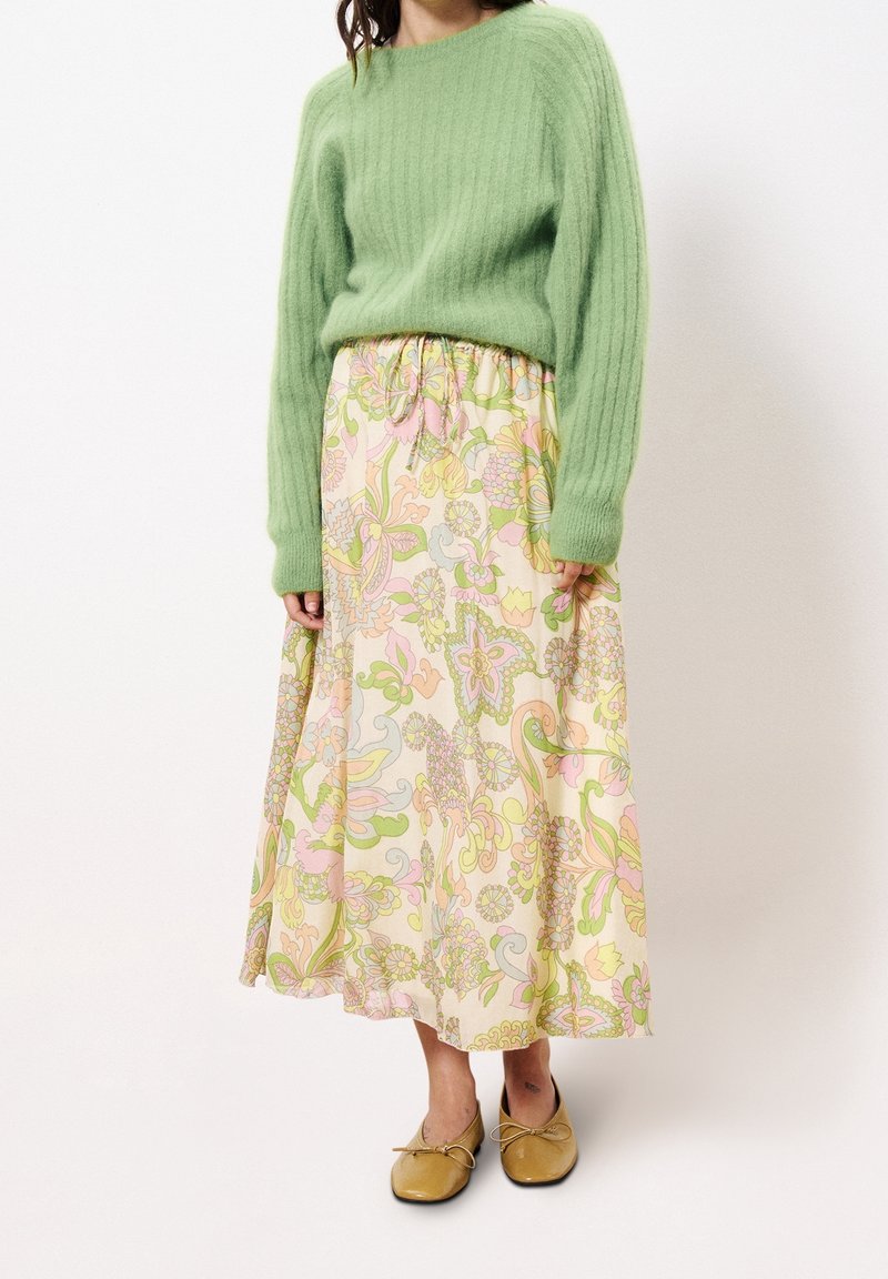 Femme portant un pull doux vert clair, une jupe midi à fleurs pastel, et des ballerines jaune moutarde, debout devant un fond blanc uni.