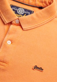 Polo orange en tissu texturé, avec un col, deux boutons et un petit logo noir "Superdry" sur la poitrine.