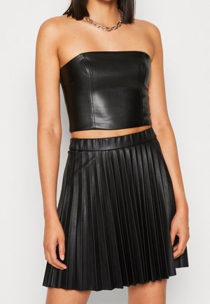 Femme portant un crop top en cuir noir sans bretelles et une jupe plissée en cuir noir assortie, accessoirisée d'un collier chaîne argenté épais.