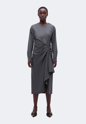 Femme portant une robe midi grise à manches longues avec des boutons asymétriques et un volant, associée à des talons noirs à bouts pointus.
