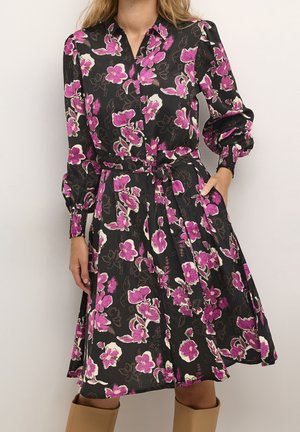 Sort kjole med lange ærmer med pink og hvidt blomsterprint, knapper foran, bælte i taljen og manchetter på ærmerne, båret med brune støvler.