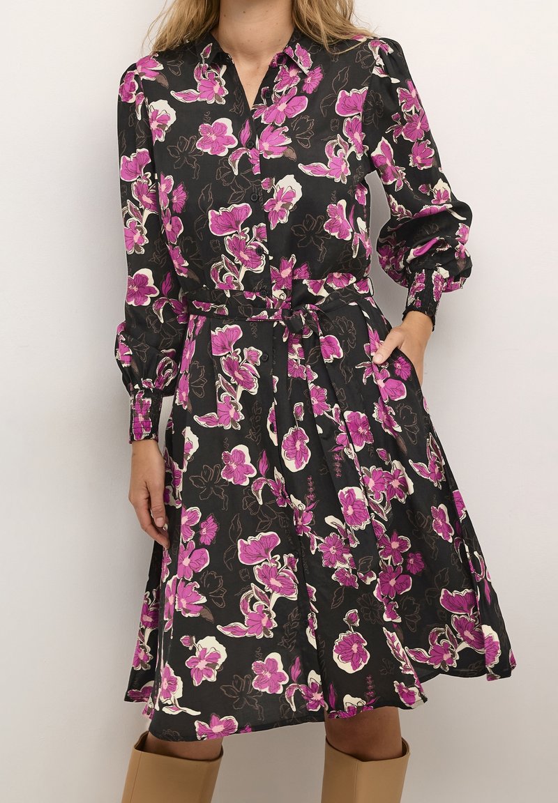 Robe noire à manches longues avec imprimé floral rose et blanc, boutonnée à l’avant, taille ceinturée et manches munies de poignets, portée avec des bottes marron.