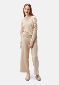 Beige stickad tröja i kombination med vida beige byxor. Mjuk textur, ribbade ärmslut och fåll. Modellen bär beige stövlar med minimalistisk design.