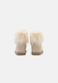Beige Wildleder-Stiefeletten mit einem weichen, flauschigen Kragen aus cremefarbigem Fell und einer flachen, tanfarbenen Gummisohle, die über glatte, strukturierte Oberflächen verfügt.